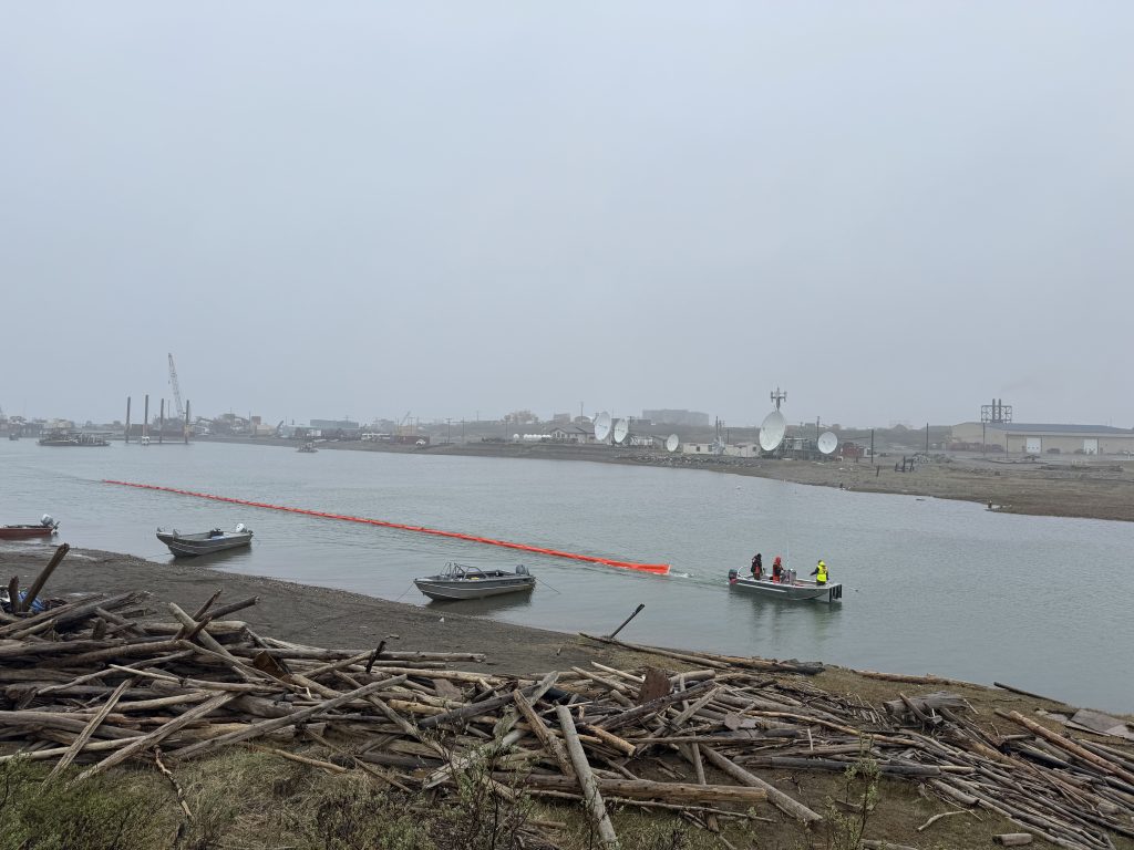 IMG_9668 - Alaska Chadux Network Harbor scene with small boats on calm water, a large pile of driftwood on the shore in the foreground, and industrial buildings in a hazy distance under a gray sky.